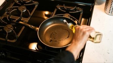 Cooking oil being poured into a black non-stick pan resting on a stovetop burner | ©Image Credit: Pexels / RDNE Stock project
