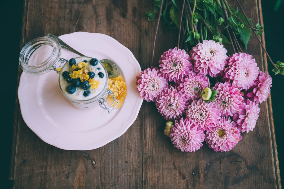 A jar of yogurt topped with blueberries and yellow garnish placed alongside a bouquet of pink flowers | ©Image Credit: Unsplash / Alisa Anton