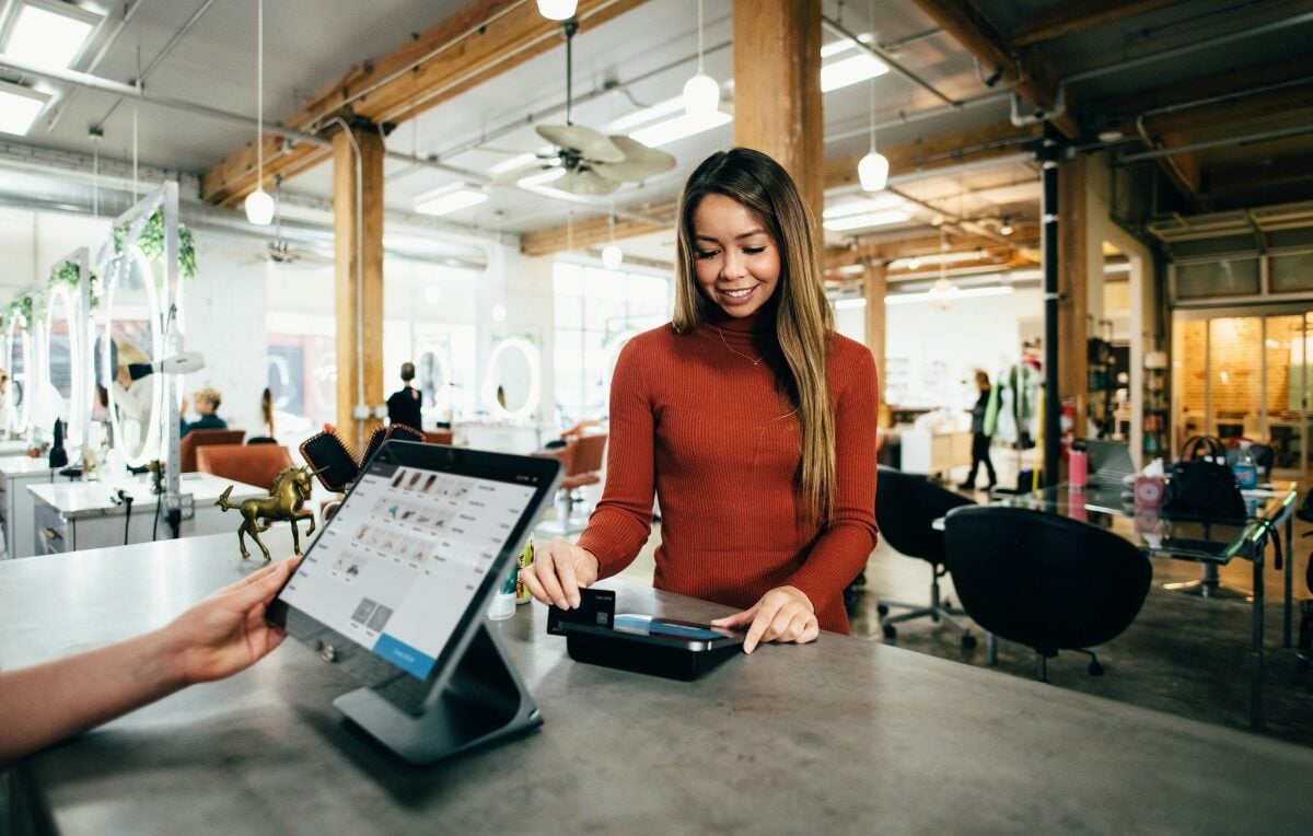 A staff member holding a tablet displaying the transaction details across the counter from a customer in a salon | ©Image Credit: Unsplash / Blake Wisz