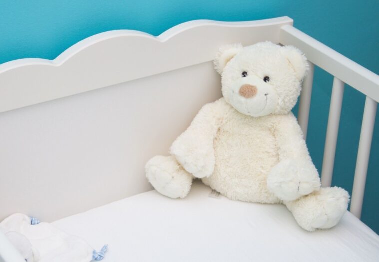 A white teddy bear sitting inside a baby's crib against a blue wall | ©Image Credit: Pixnio