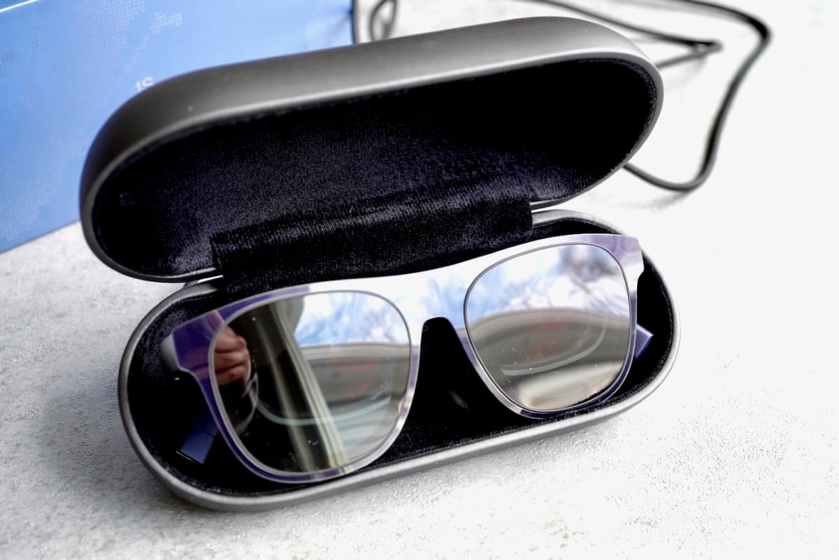 The XREAL 1S glasses neatly stored in their premium matte black hard carrying case, with the dark-tinted lenses reflecting the blue retail box and surroundings