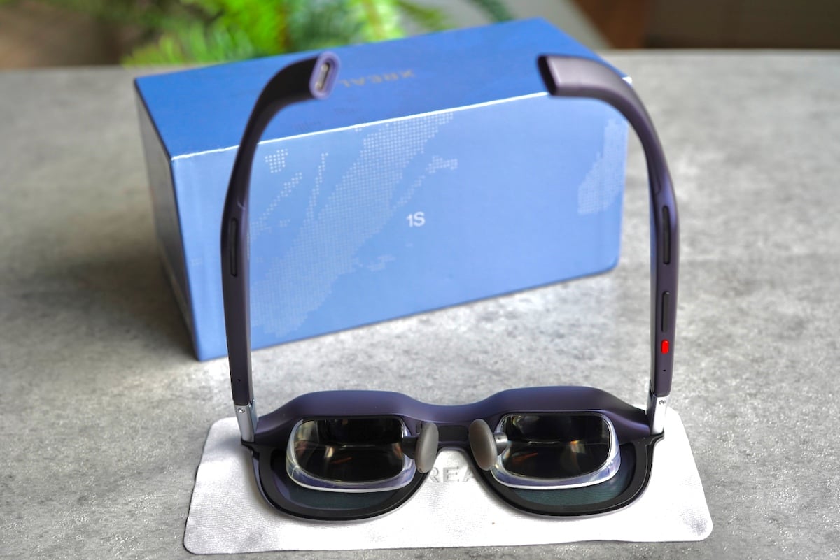 The XREAL 1S glasses propped up in front of their blue retail box, clearly displaying the red status/control button, USB-C port, and curved purple temples