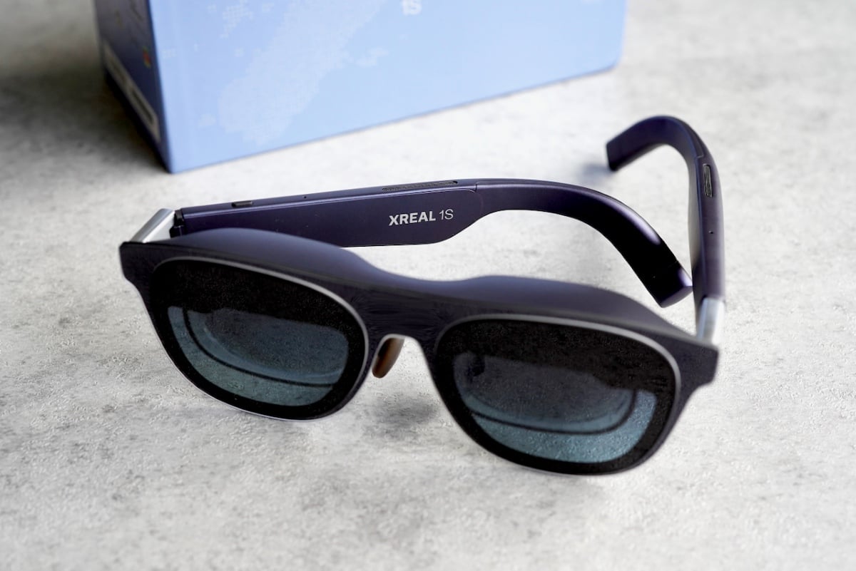 The XREAL 1S spatial display glasses resting on a light surface, showcasing their full front view with dark tinted lenses, matte black frame, and distinctive purple temples branded "XREAL 1S