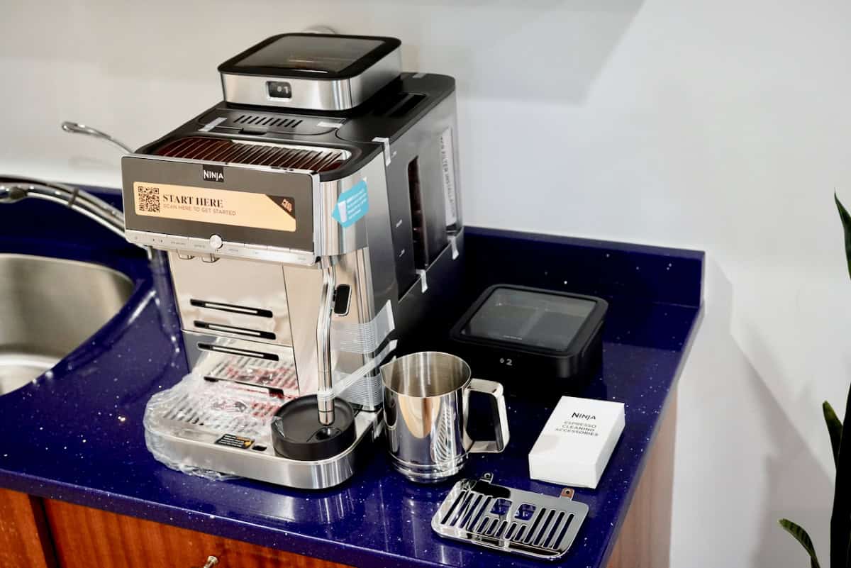 Unboxed Ninja AutoBarista Pro on a kitchen counter with protective tape and accessories, including a milk pitcher and cleaning kit