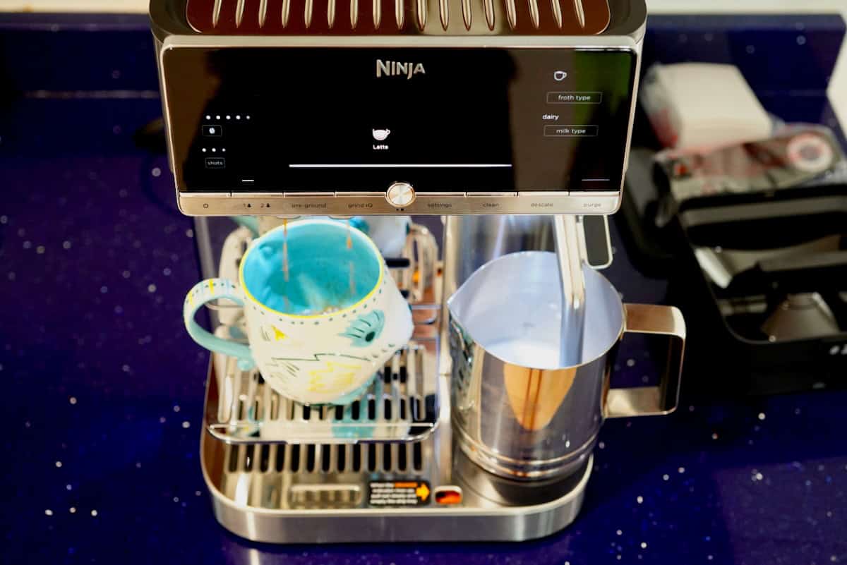Top-down view of the Ninja AutoBarista Pro actively dispensing coffee into a ceramic mug alongside a stainless steel milk frothing pitcher.